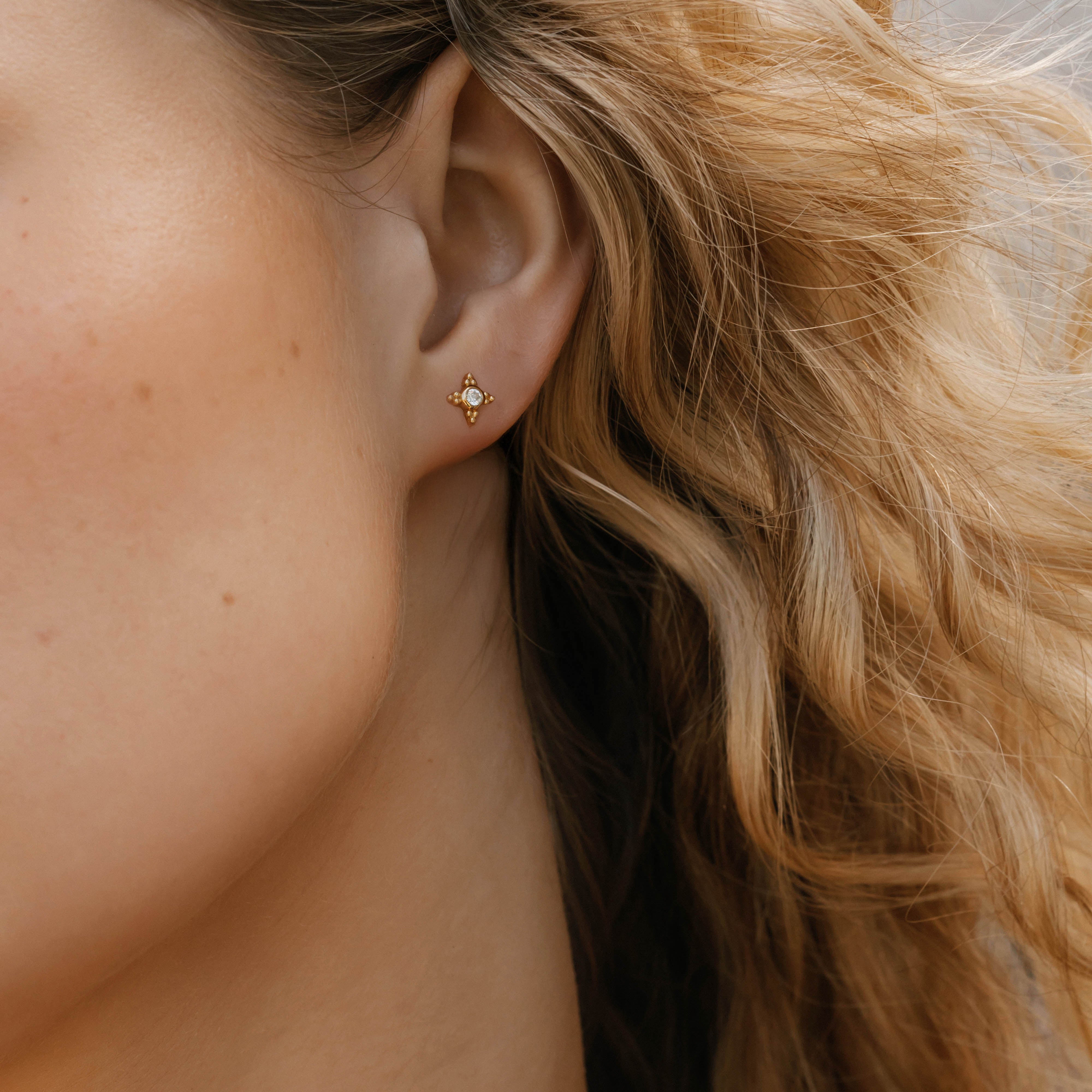 A close-up shows a woman's ear wearing LLUME Jewelry's Myra Studs—delicate earrings with a tiny central gemstone. Her wavy blonde hair frames her naturally toned skin, while a softly blurred background accentuates the jewelry.