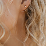 Close-up of a person's ear with wavy blonde hair, wearing LLUME Jewelry's Myra Studs—small gold star-shaped earrings that make an elegant addition to any jewelry collection.