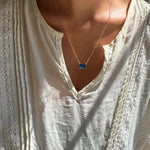 A person wearing a cream-colored, lacy blouse and LLUME Jewelry's Seacove Necklace—a silver multilength chain featuring a rectangular sapphire-blue pendant—shown in natural sunlight.