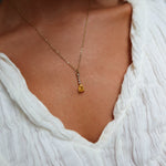 A close-up of a person in a textured white V-neck top wearing the LLUME Jewelry Sunspill Necklace, featuring a delicate gold chain and yellow teardrop pendant.
