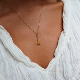 A close-up of a person in a textured white V-neck top wearing the LLUME Jewelry Sunspill Necklace, featuring a delicate gold chain and yellow teardrop pendant.