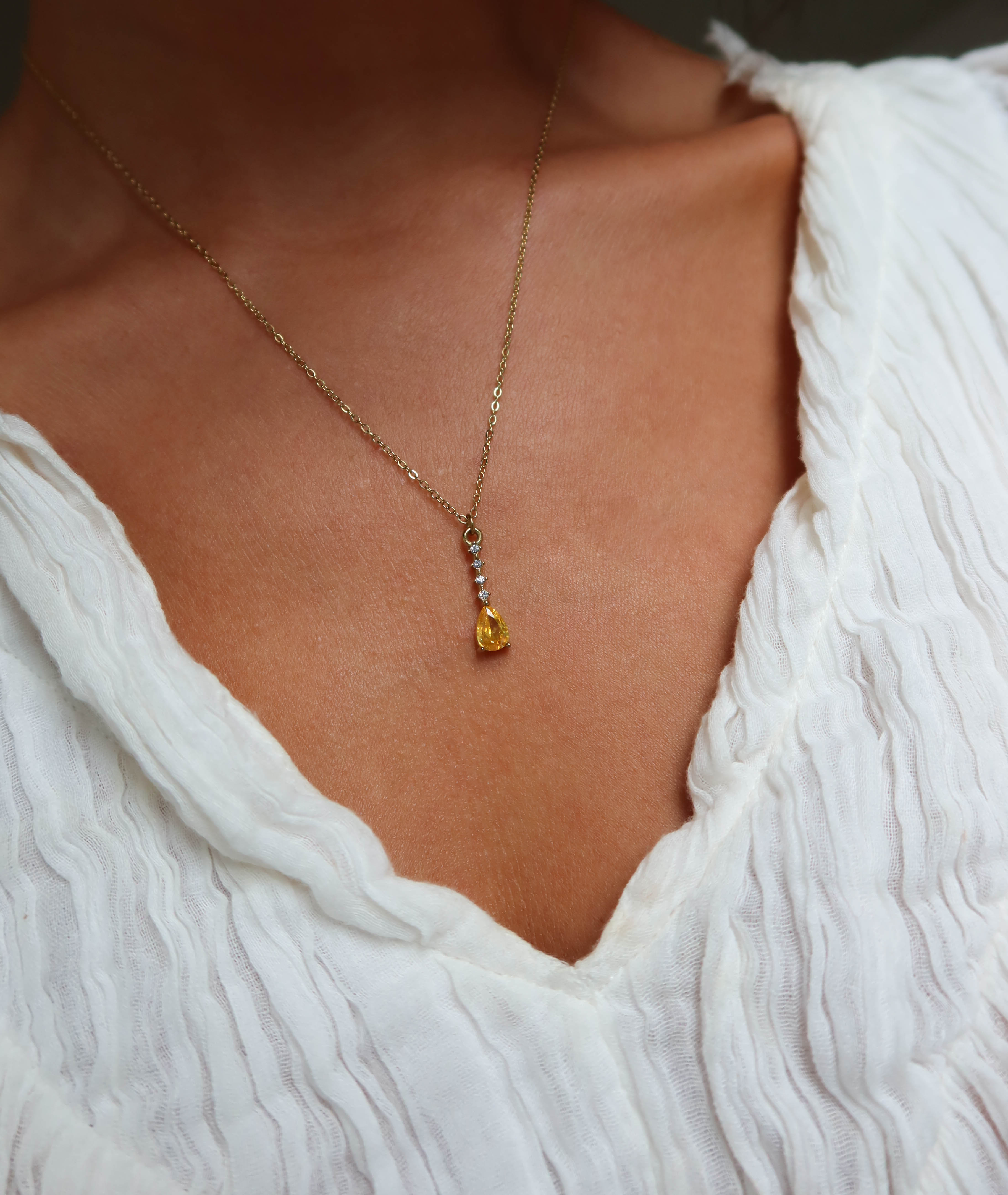 A close-up of a person in a textured white V-neck top wearing the LLUME Jewelry Sunspill Necklace, featuring a delicate gold chain and yellow teardrop pendant.