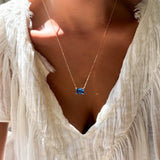A person wearing a textured white blouse and the LLUME Jewelry Seacove Necklace—a delicate multilength chain featuring a rectangular sapphire-blue pendant—photographed in natural light.