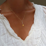 A person wearing a white textured blouse and the Sunspill Necklace by LLUME Jewelry, a delicate gold waterproof necklace with a small yellow pendant.
