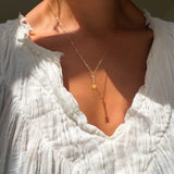 A person in a crinkled white blouse wears the LLUME Jewelry Sunspill Necklace—a delicate, waterproof piece with a small yellow pendant—shown from neck to upper chest in sunlight.