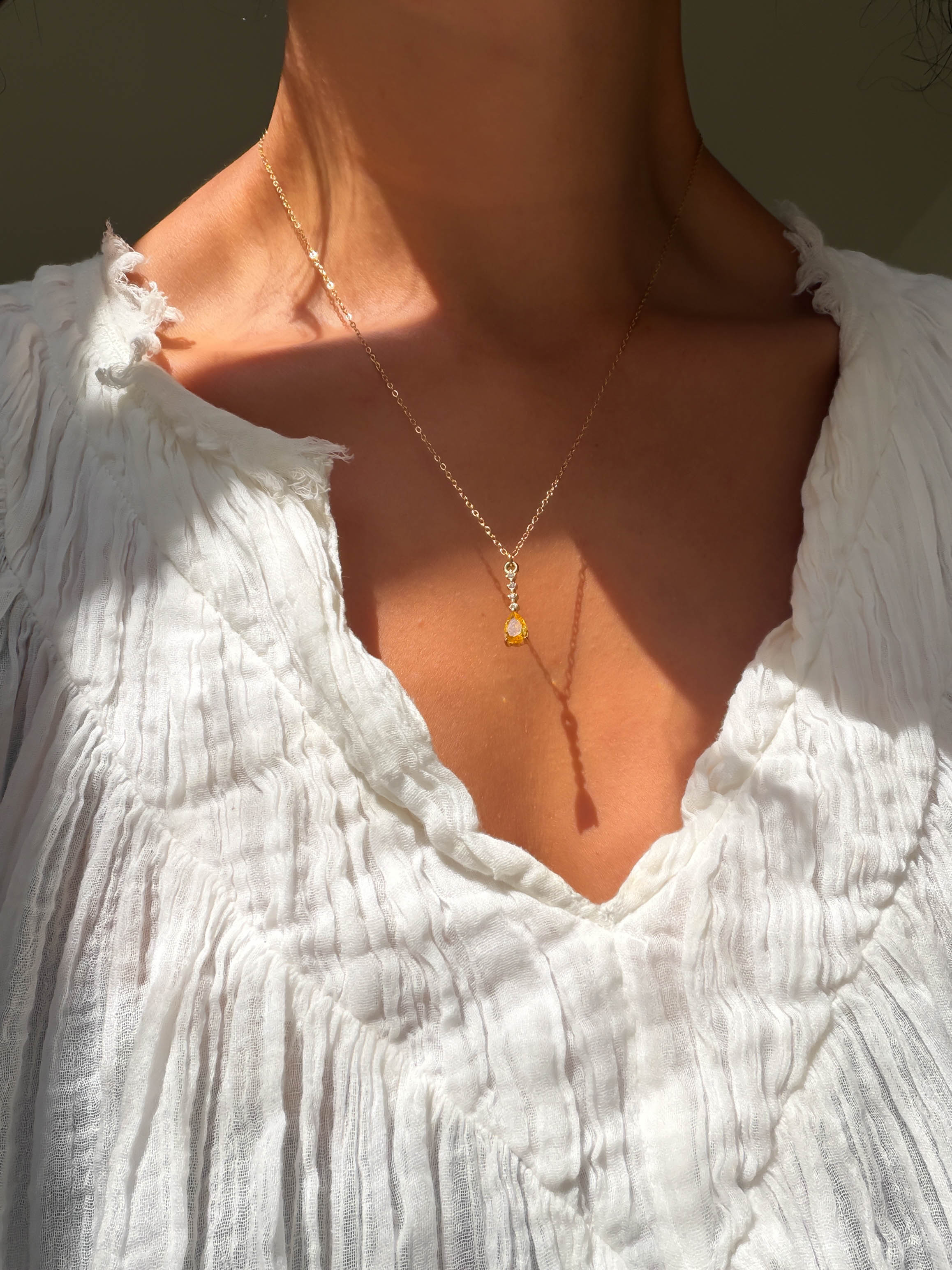 A person in a crinkled white blouse wears the LLUME Jewelry Sunspill Necklace—a delicate, waterproof piece with a small yellow pendant—shown from neck to upper chest in sunlight.