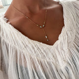 A person wearing the Ever Necklace by LLUME Jewelry, a delicate waterproof gold piece with blue and white stones, paired with a white textured v-neck blouse.