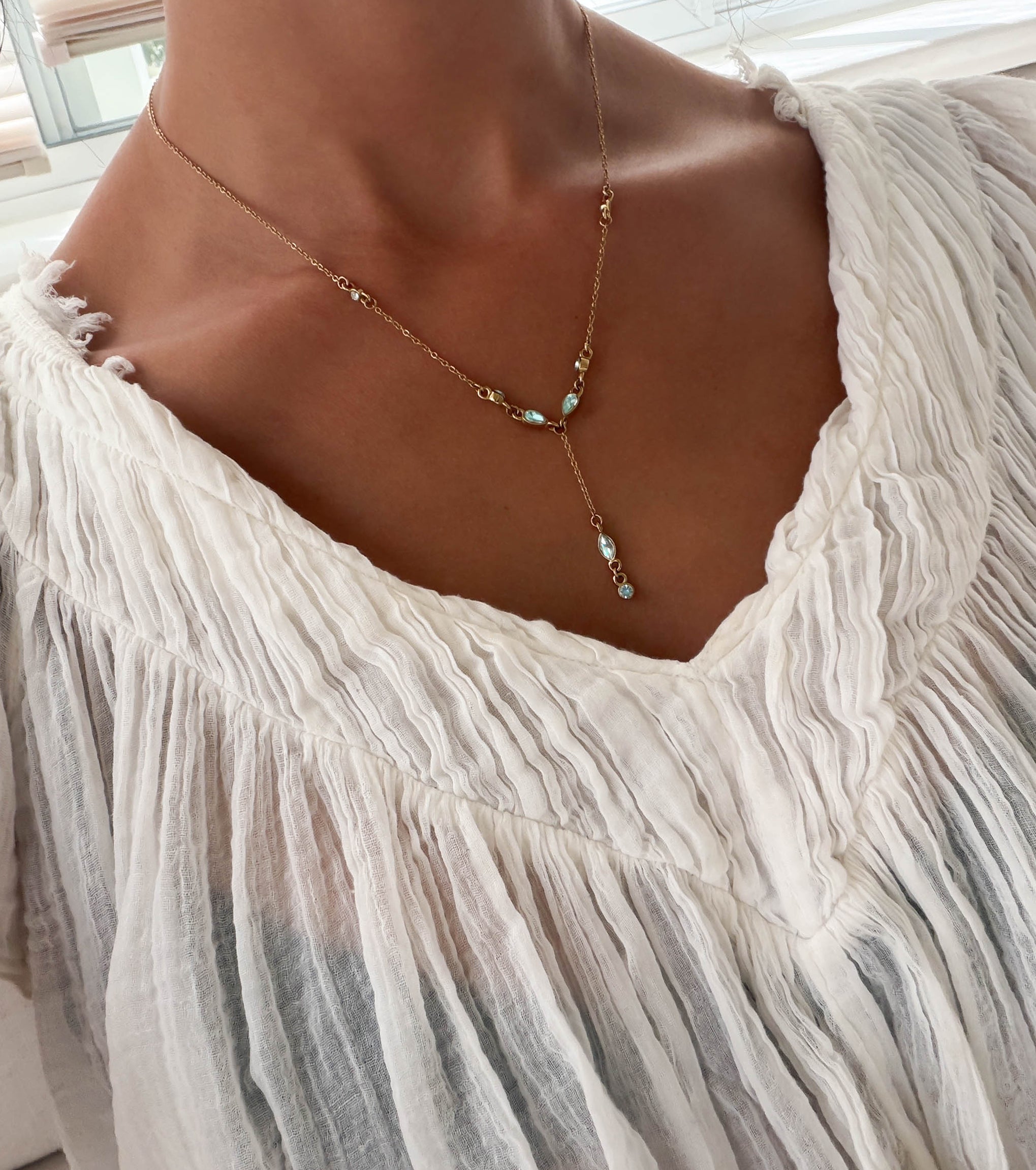 A person wearing the Ever Necklace by LLUME Jewelry, a delicate waterproof gold piece with blue and white stones, paired with a white textured v-neck blouse.