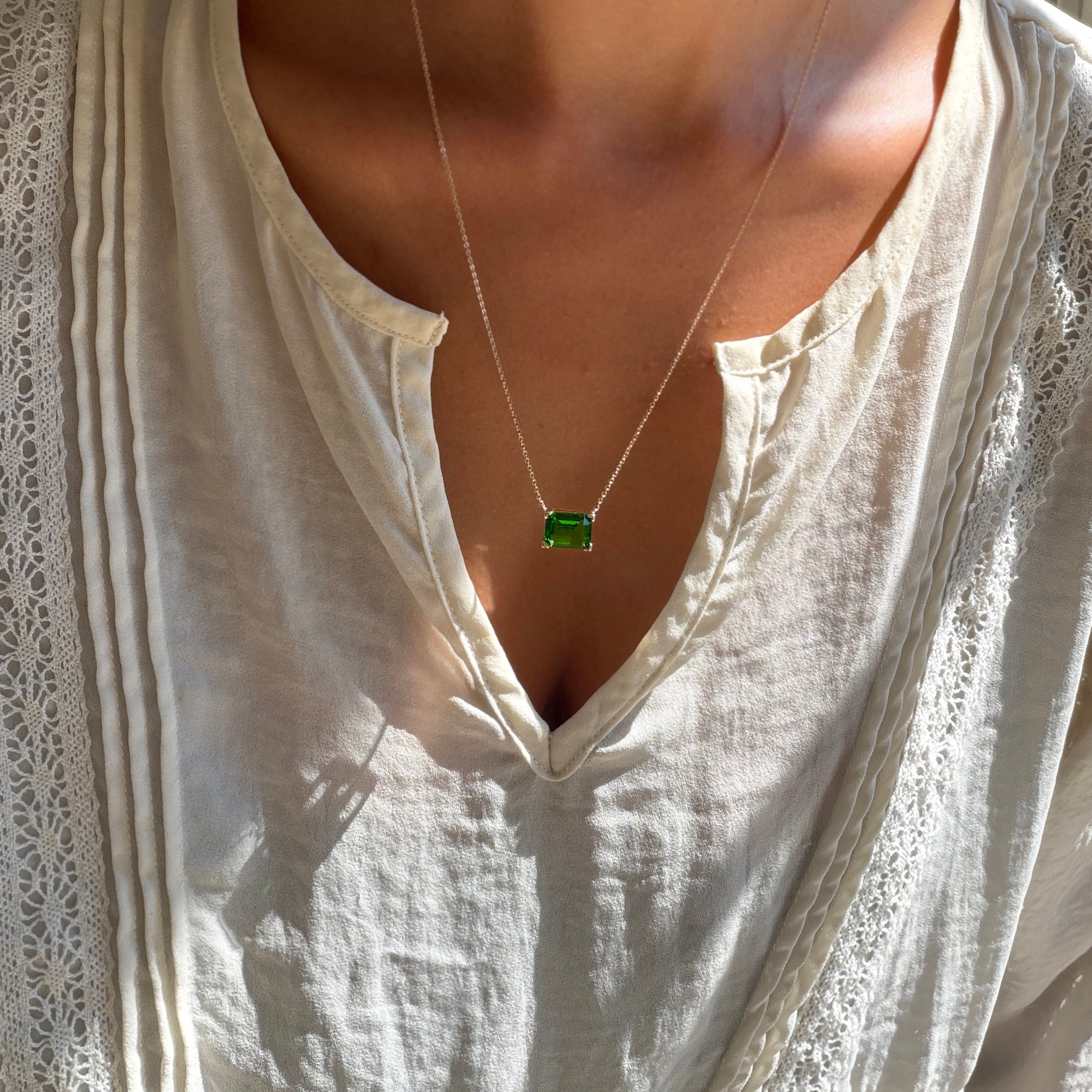 A person wears a white, textured blouse with a v-shaped neckline, accented by LLUME Jewelry’s Seagrass Necklace—a green pendant hanging from a delicate multilength chain. Sunlight casts gentle shadows across the blouse.