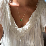 A person wearing a textured white blouse and the LLUME Jewelry Seagrass Necklace—a delicate gold-plated multilength chain with a green rectangular gemstone pendant—shown in natural sunlight.