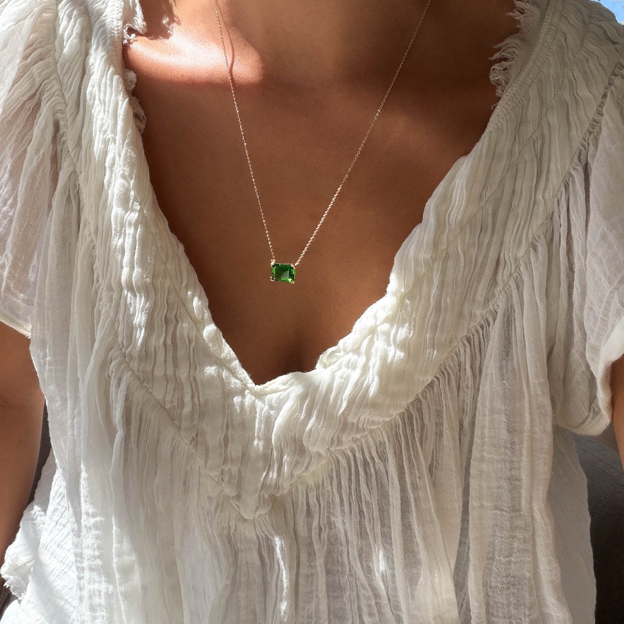 A person wearing a textured white blouse and the LLUME Jewelry Seagrass Necklace—a delicate gold-plated multilength chain with a green rectangular gemstone pendant—shown in natural sunlight.