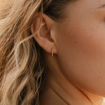 A close-up shows wavy blonde hair and an ear adorned with LLUME Jewelry's Vacation Capsule Set—featuring a small gold hoop earring and delicate chain necklaces for an elegant, stackable look.