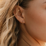 A close-up shows wavy blonde hair and an ear adorned with LLUME Jewelry's Vacation Capsule Set—featuring a small gold hoop earring and delicate chain necklaces for an elegant, stackable look.