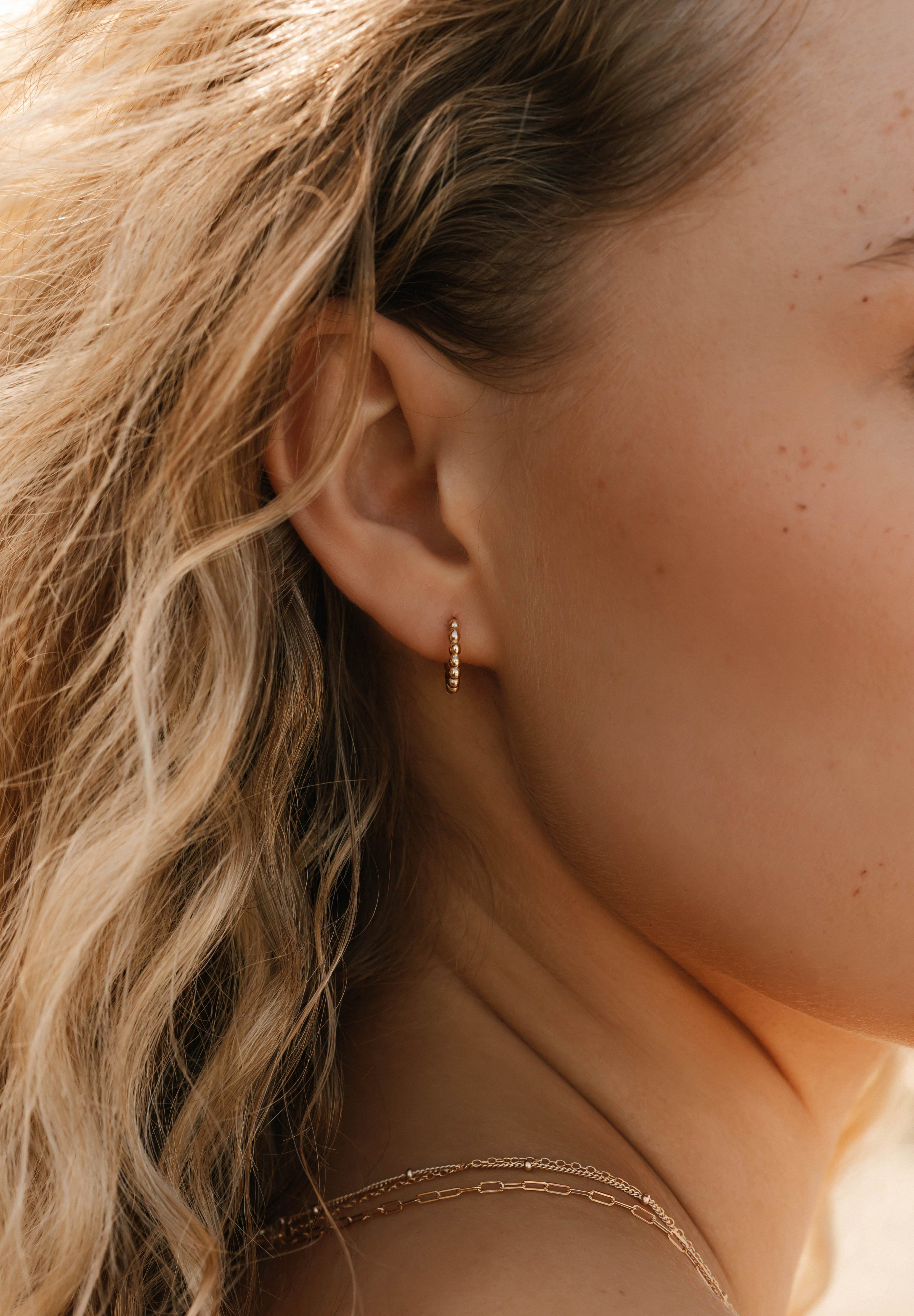 A close-up shows wavy blonde hair and an ear adorned with LLUME Jewelry's Vacation Capsule Set—featuring a small gold hoop earring and delicate chain necklaces for an elegant, stackable look.