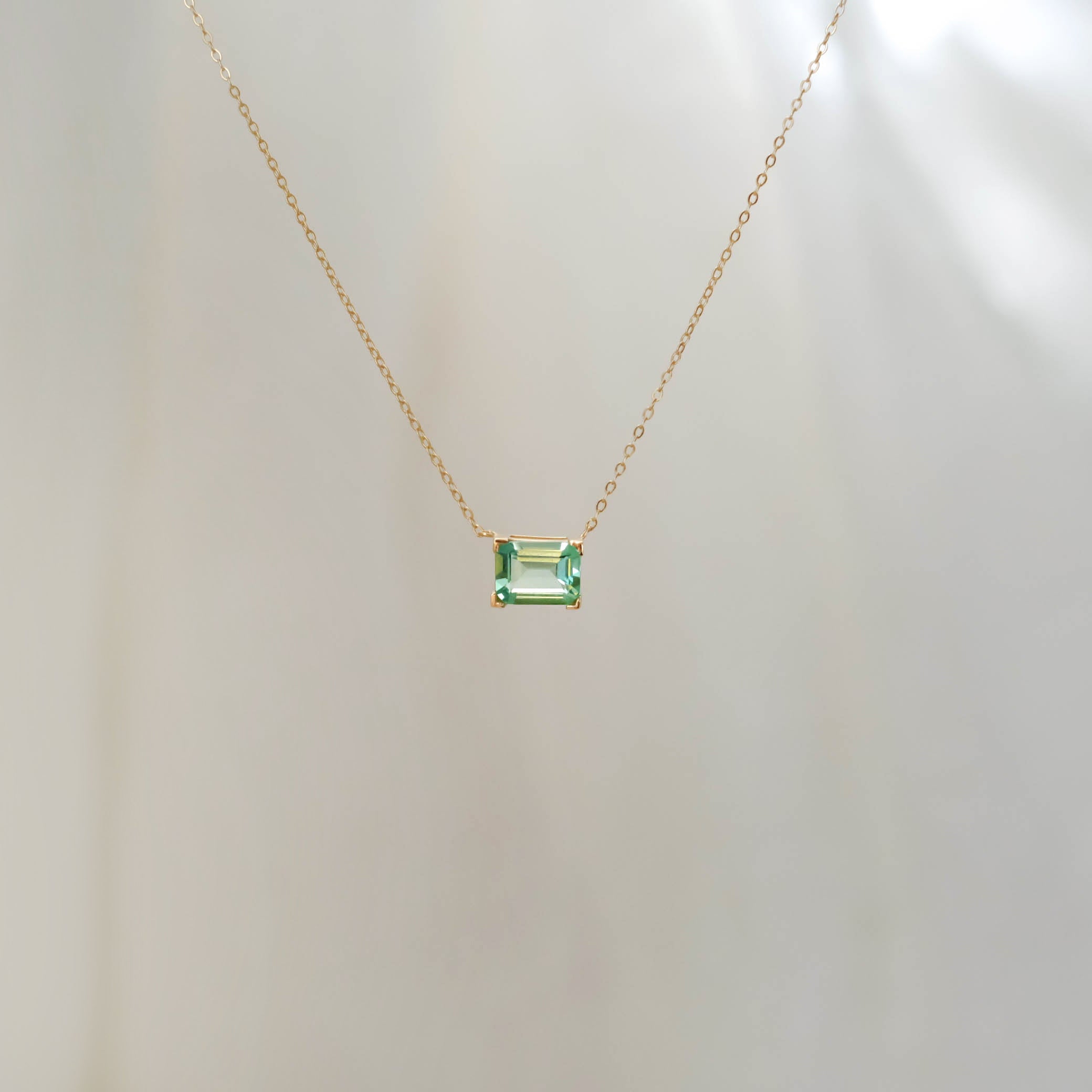 The Seafoam Glass Necklace by LLUME Jewelry features a delicate gold chain with a rectangular green glass pendant, evoking coastal magic against a light neutral background.