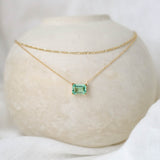 The LLUME Jewelry Seafoam Daily Set, featuring a delicate chain with a rectangular green gemstone pendant, is draped over a smooth beige vase against a soft white fabric background.