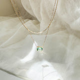 The LLUME Jewelry Seafoam Daily Set, featuring a delicate gold chain with a rectangular green gemstone pendant, rests on sheer white fabric, softly illuminated by natural light.