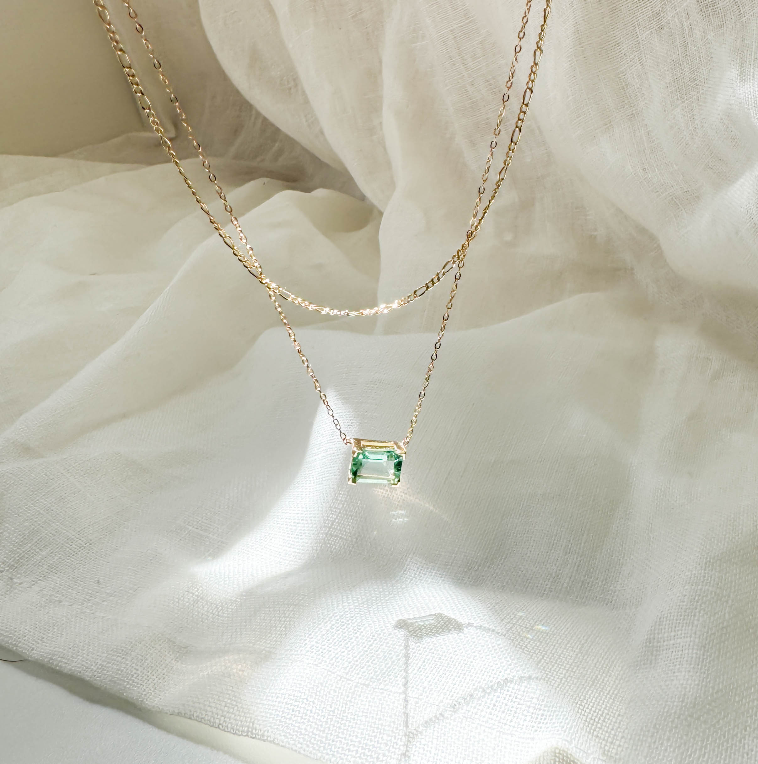 The LLUME Jewelry Seafoam Daily Set, featuring a delicate gold chain with a rectangular green gemstone pendant, rests on sheer white fabric, softly illuminated by natural light.