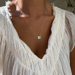 A person wears a sheer, textured white blouse with a deep V-neck, styled with LLUME Jewelry’s Seafoam Glass Necklace—a delicate gold piece featuring a rectangular green gemstone pendant that evokes coastal magic.