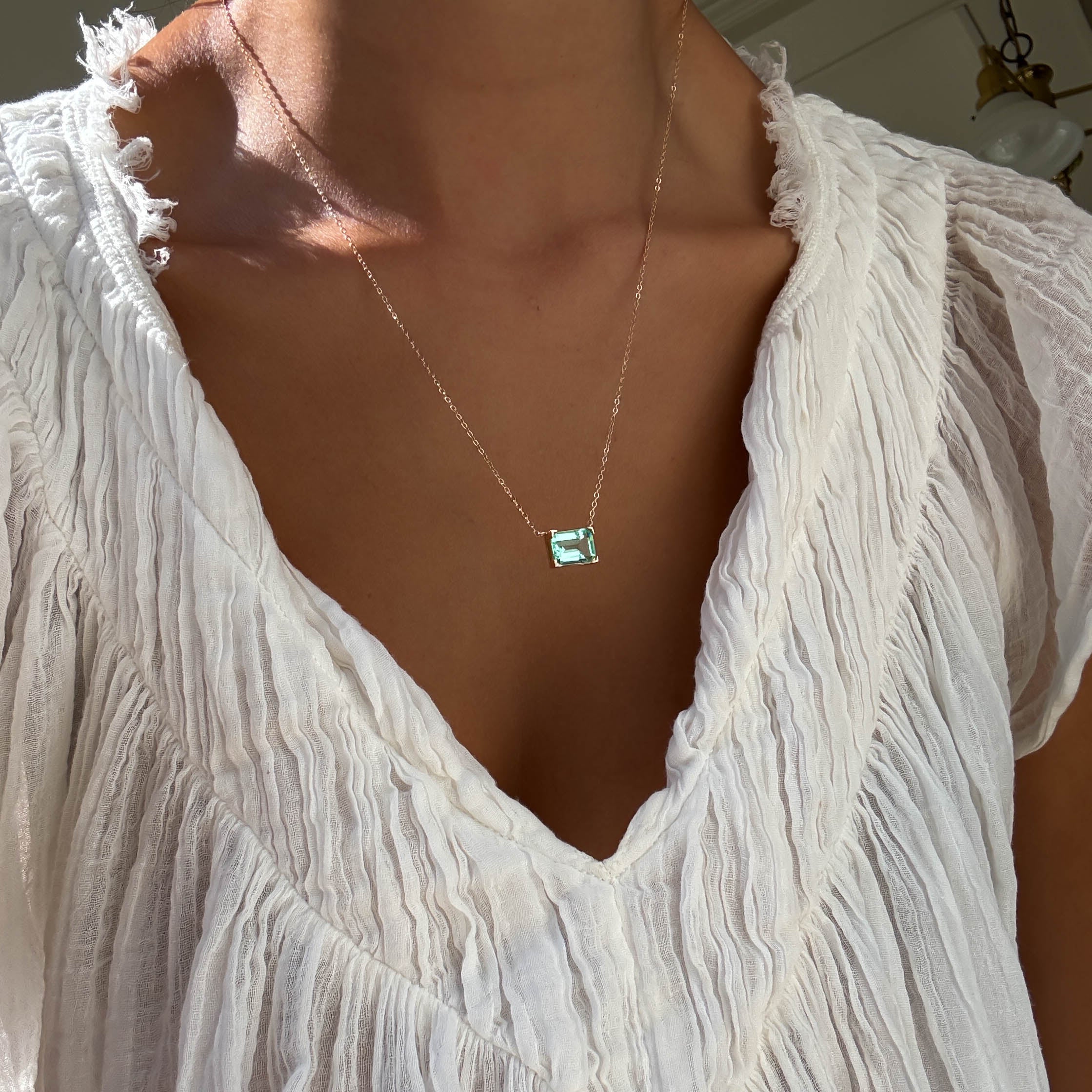A person wears a sheer, textured white blouse with a deep V-neck, styled with LLUME Jewelry’s Seafoam Glass Necklace—a delicate gold piece featuring a rectangular green gemstone pendant that evokes coastal magic.