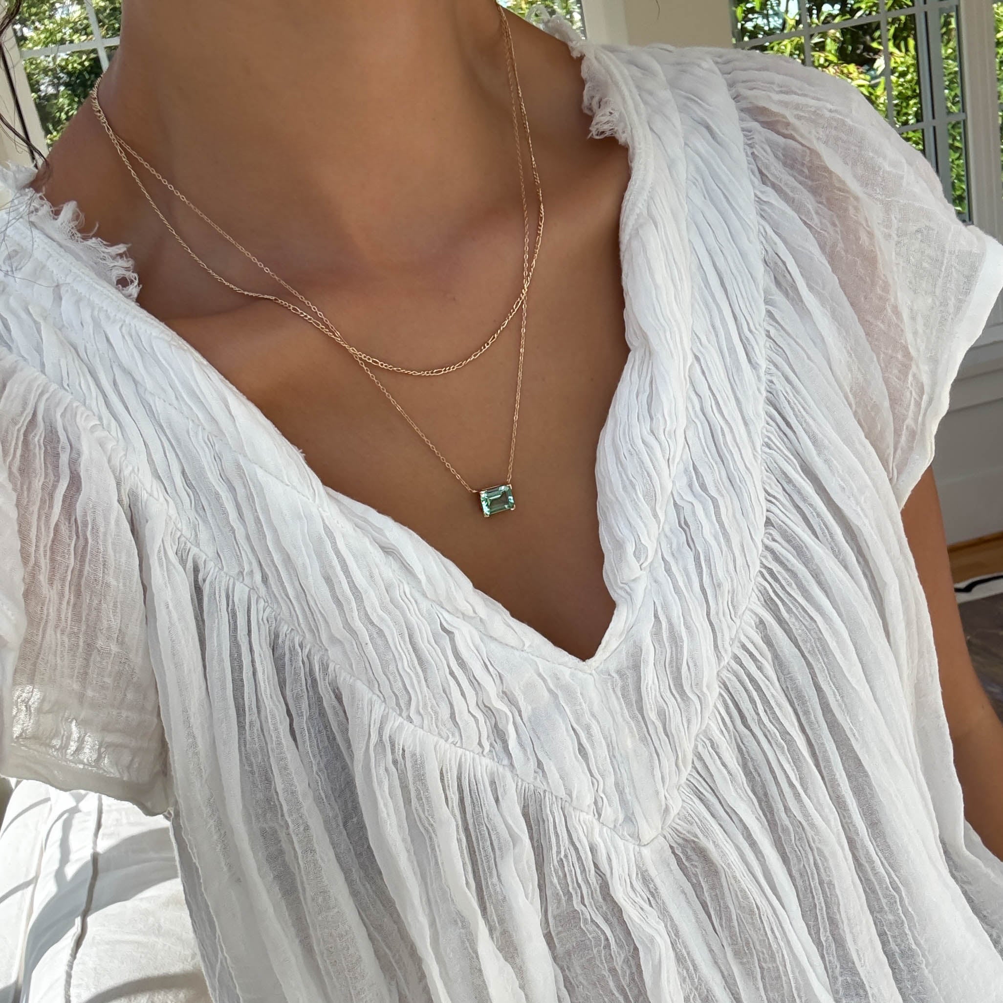 In a sunlit setting, someone wears a sheer white blouse with a deep V-neck, styled with the LLUME Jewelry Seafoam Daily Set—a delicate necklace set featuring the signature green pendant.