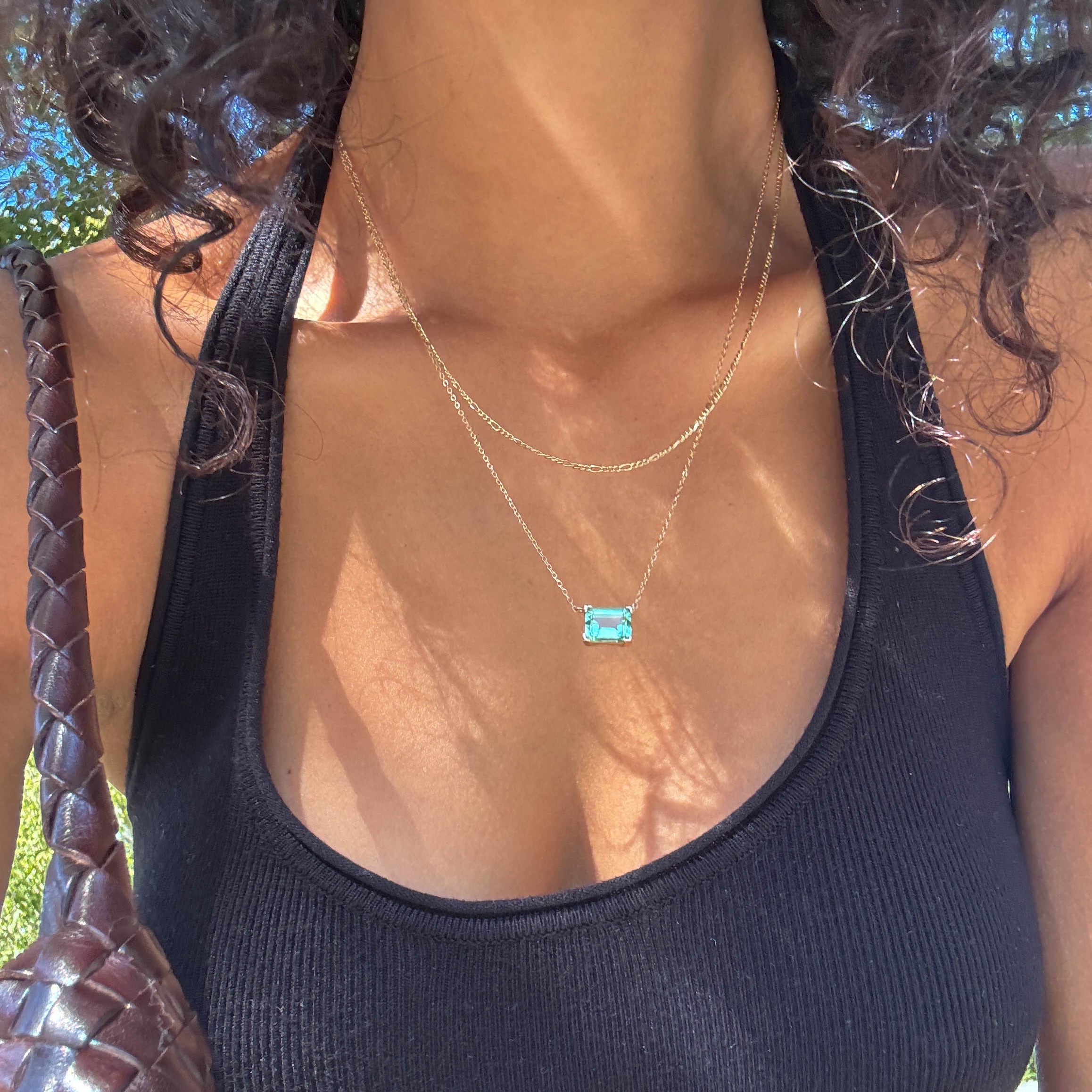 A person with curly hair wears a black ribbed halter top and carries a woven brown bag. Sunlight casts shadows across their chest, highlighting the LLUME Jewelry Seafoam Daily Set around their neckline.