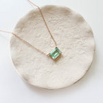 The Seafoam Glass Necklace by LLUME Jewelry features a delicate gold chain and a rectangular light green seafoam glass pendant, displayed on an off-white ceramic dish for a touch of coastal elegance.