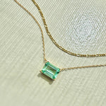 The LLUME Jewelry Seafoam Daily Set, featuring a gold chain necklace with a thin strand and a square-cut green gemstone pendant, rests on a textured light surface in bright sunlight.