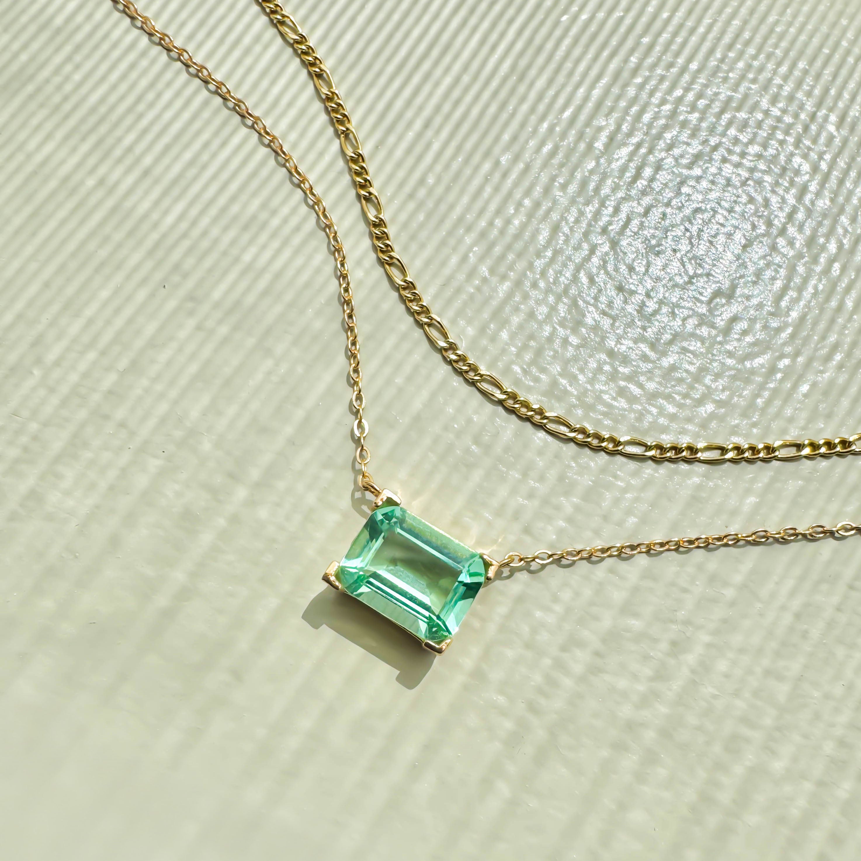 The LLUME Jewelry Seafoam Daily Set, featuring a gold chain necklace with a thin strand and a square-cut green gemstone pendant, rests on a textured light surface in bright sunlight.
