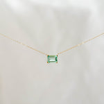 The Seafoam Glass Necklace by LLUME Jewelry features a delicate gold chain and a small rectangular green pendant, evoking coastal magic against a soft, light background.