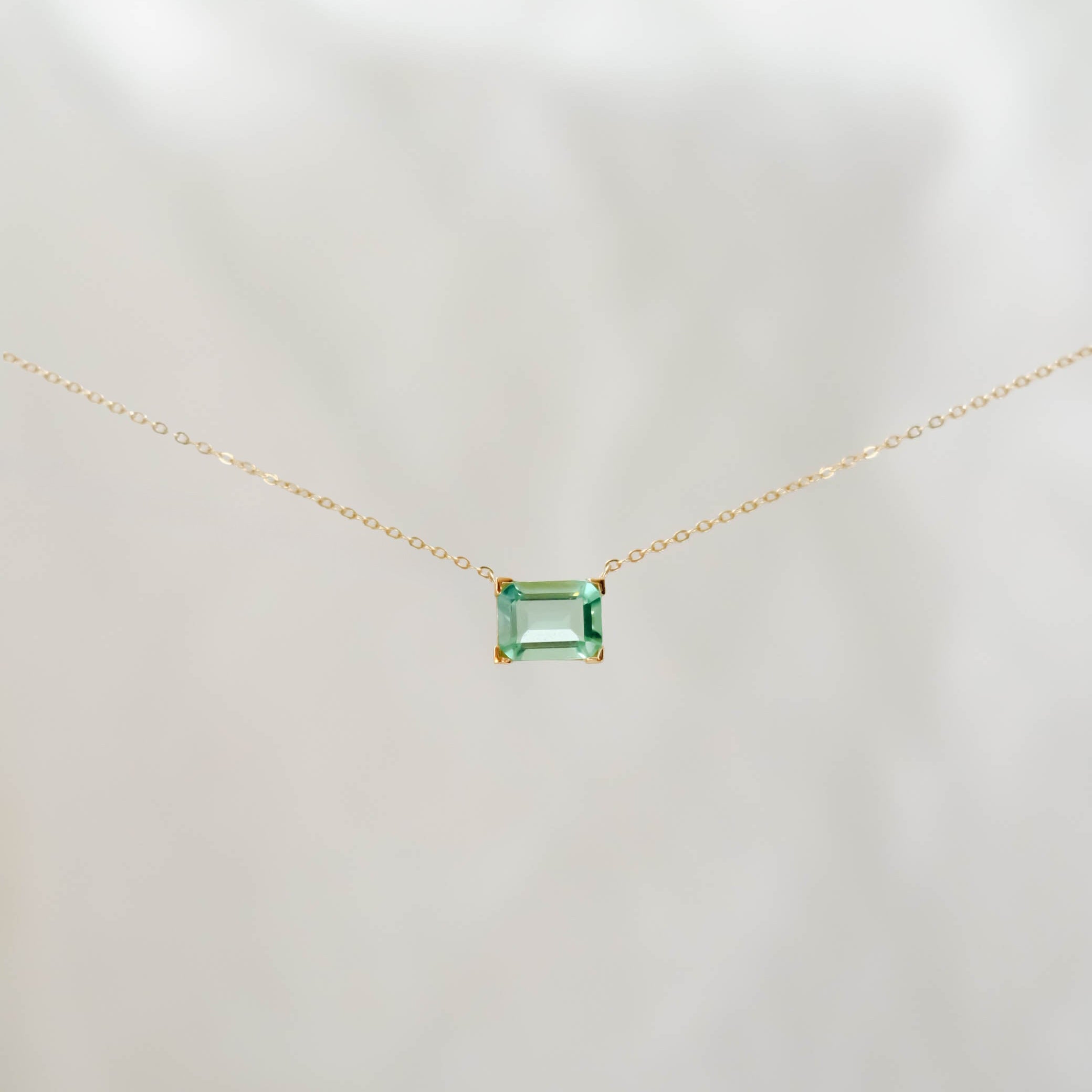The Seafoam Glass Necklace by LLUME Jewelry features a delicate gold chain and a small rectangular green pendant, evoking coastal magic against a soft, light background.