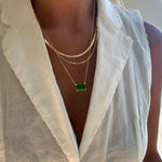 Close-up of a person wearing a sleeveless white collared blouse, styled with the LLUME Jewelry Seagrass Favorites Set: three layered gold necklaces, including one with a green rectangular pendant and another featuring an elegant paperclip chain.