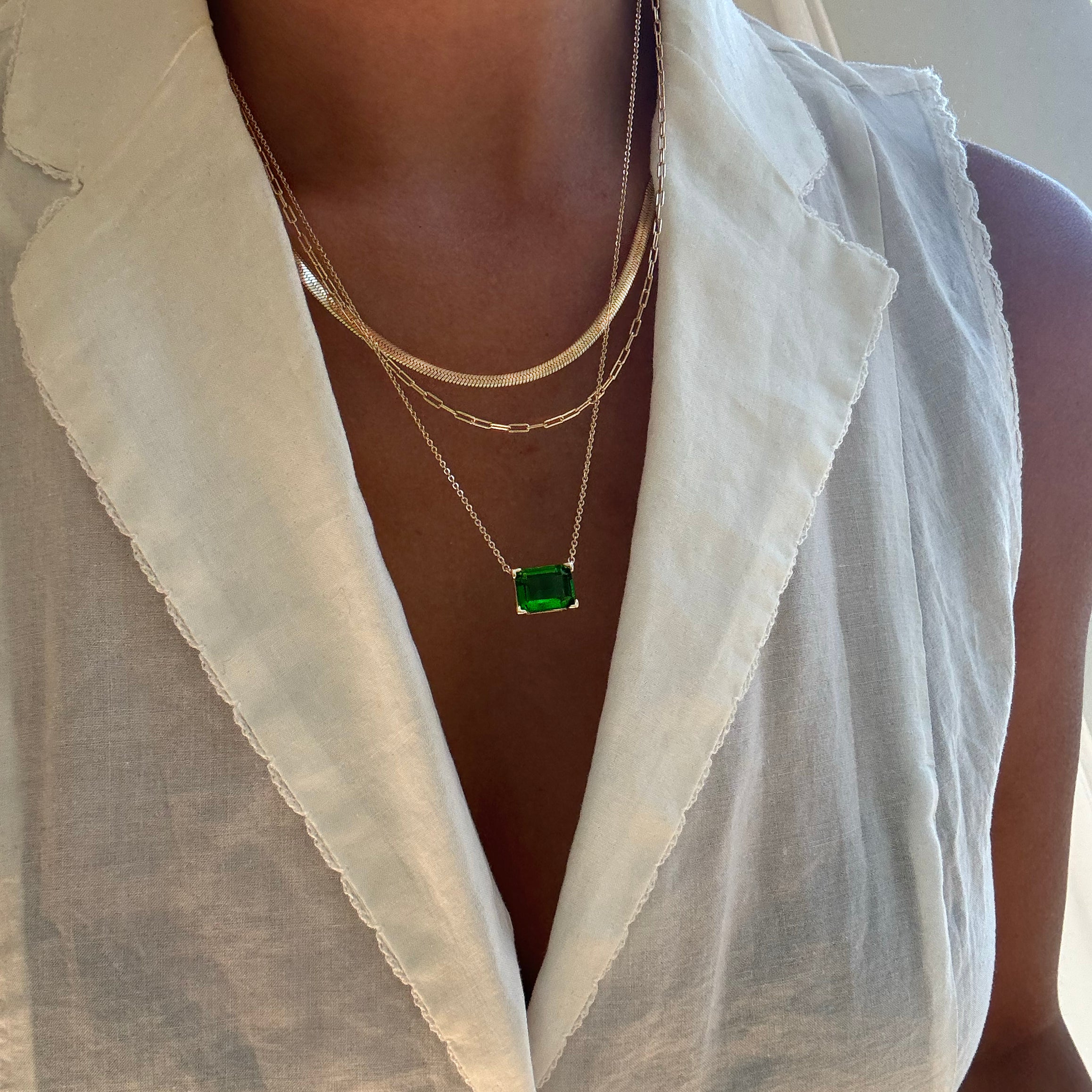 Close-up of a person wearing a sleeveless white collared blouse, styled with the LLUME Jewelry Seagrass Favorites Set: three layered gold necklaces, including one with a green rectangular pendant and another featuring an elegant paperclip chain.