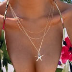 A close-up of a person in a floral bikini top wearing LLUME Jewelry's Starfish Favorites Set layered gold necklaces, including a golden hour chain and starfish necklace, against a sunny outdoor backdrop.