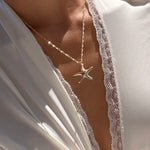 A person wearing a silky white robe with lace trim showcases the LLUME Jewelry Starfish Necklace, its seaside charm glinting in the sunlight.