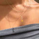 A person wears LLUME Jewelry’s Sunspill Daily Set: a grey off-shoulder top pairs with two delicate gold chains, one featuring the signature Sunspill Necklace and its teardrop yellow pendant glowing in natural light.