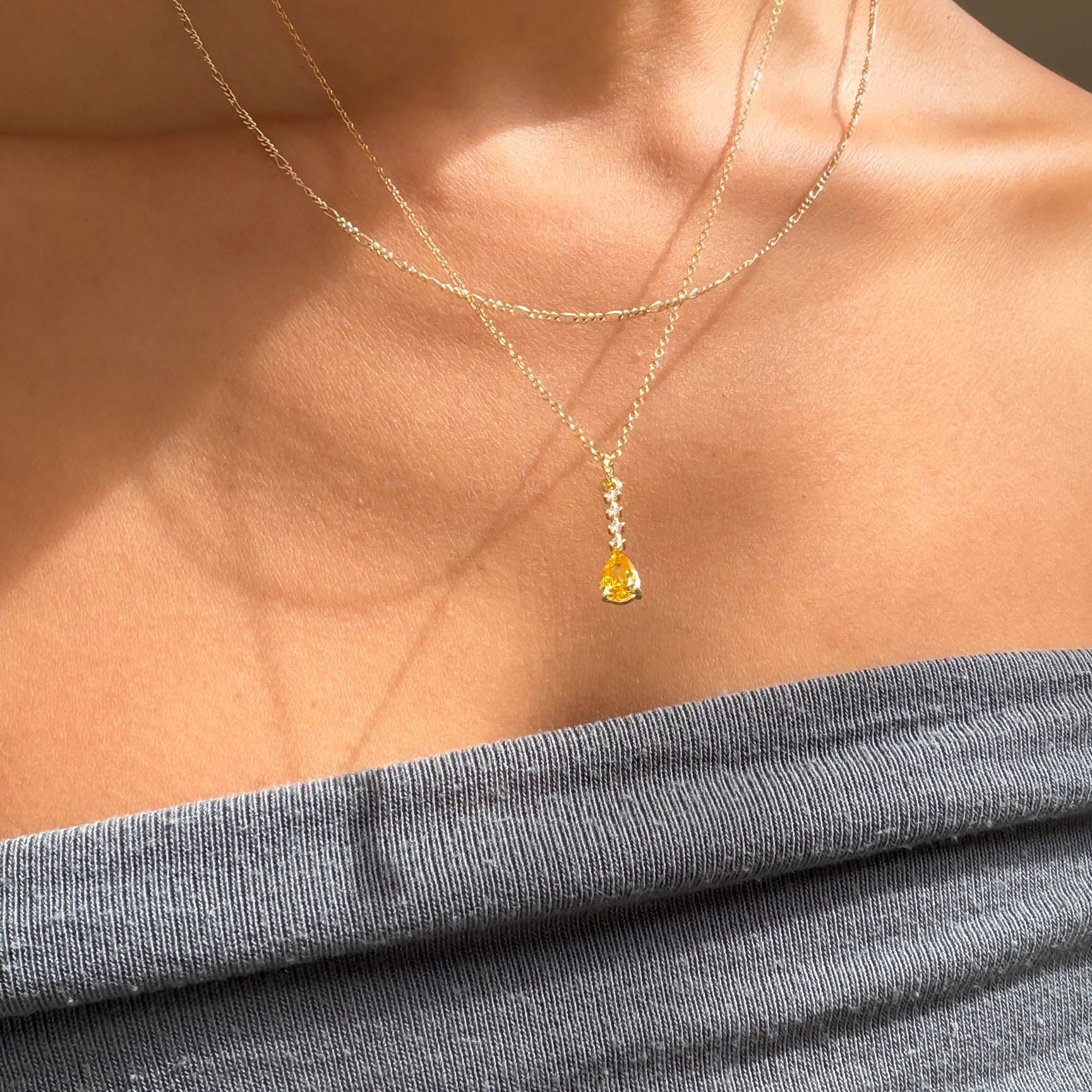 A person wears LLUME Jewelry’s Sunspill Daily Set: a grey off-shoulder top pairs with two delicate gold chains, one featuring the signature Sunspill Necklace and its teardrop yellow pendant glowing in natural light.