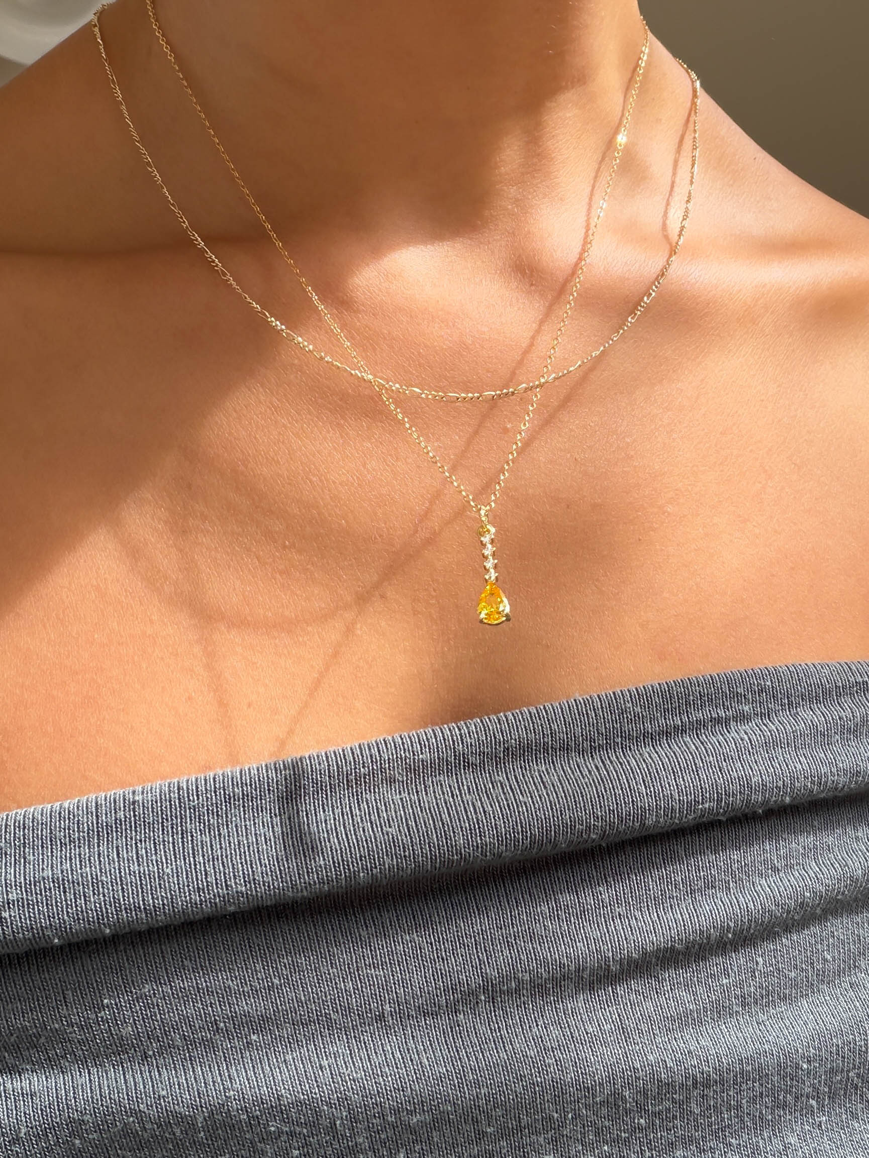 A person wears LLUME Jewelry’s Sunspill Daily Set: a grey off-shoulder top pairs with two delicate gold chains, one featuring the signature Sunspill Necklace and its teardrop yellow pendant glowing in natural light.
