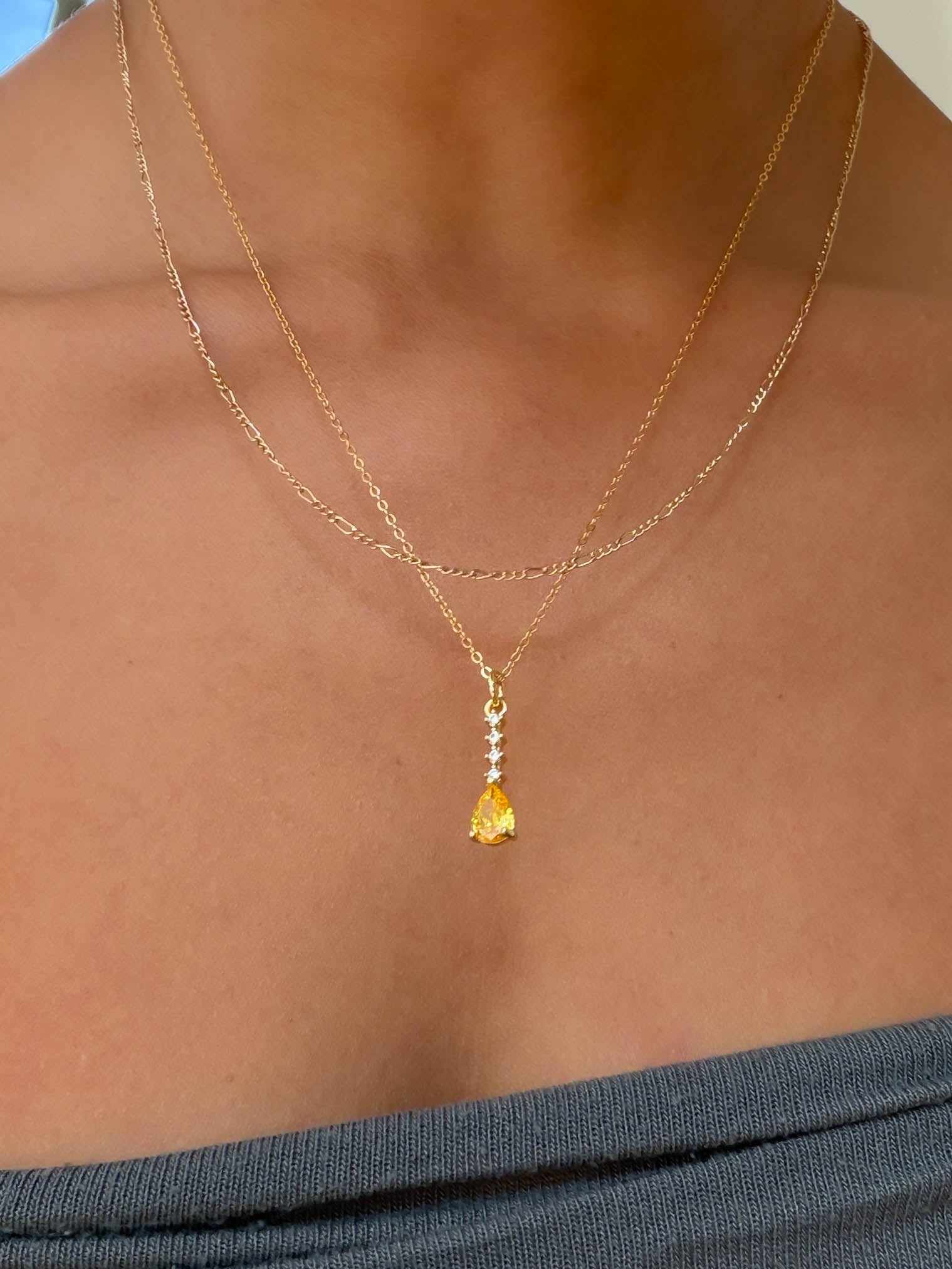 A person wears the LLUME Jewelry Sunspill Daily Set, featuring a necklace with a yellow teardrop pendant and small stone accents, paired with a grey strapless top.