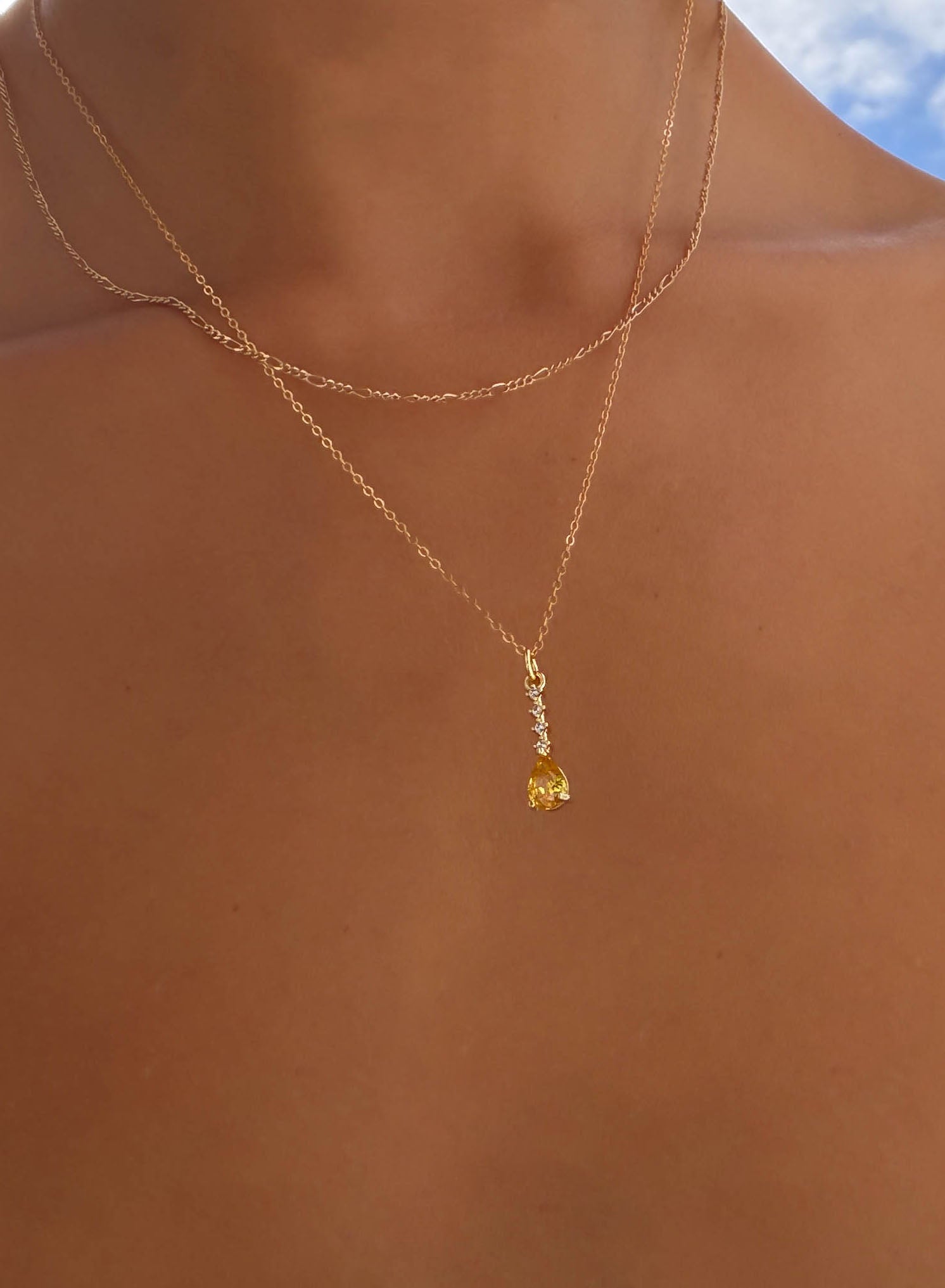 A close-up of a person’s neck wearing LLUME Jewelry’s Sunspill Daily Set—two delicate gold chains, one with a small yellow pendant—set against a blurred outdoor background.