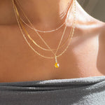 A person wears the LLUME Jewelry Sunspill Solstice Set, featuring layered gold necklaces and a small yellow gemstone pendant, paired with an off-the-shoulder gray top as sunlight highlights their collarbones and skin.