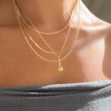 A person wears the LLUME Jewelry Sunspill Solstice Set, featuring layered gold necklaces and a small yellow gemstone pendant, paired with an off-the-shoulder gray top as sunlight highlights their collarbones and skin.