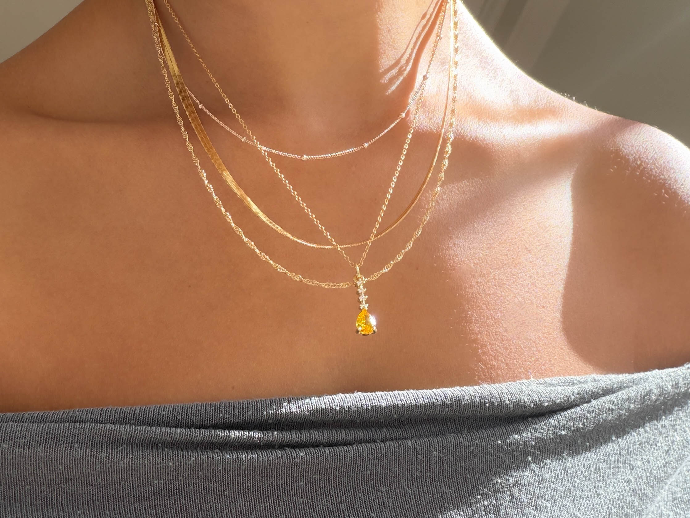 A person wears the LLUME Jewelry Sunspill Solstice Set, featuring layered gold necklaces and a small yellow gemstone pendant, paired with an off-the-shoulder gray top as sunlight highlights their collarbones and skin.