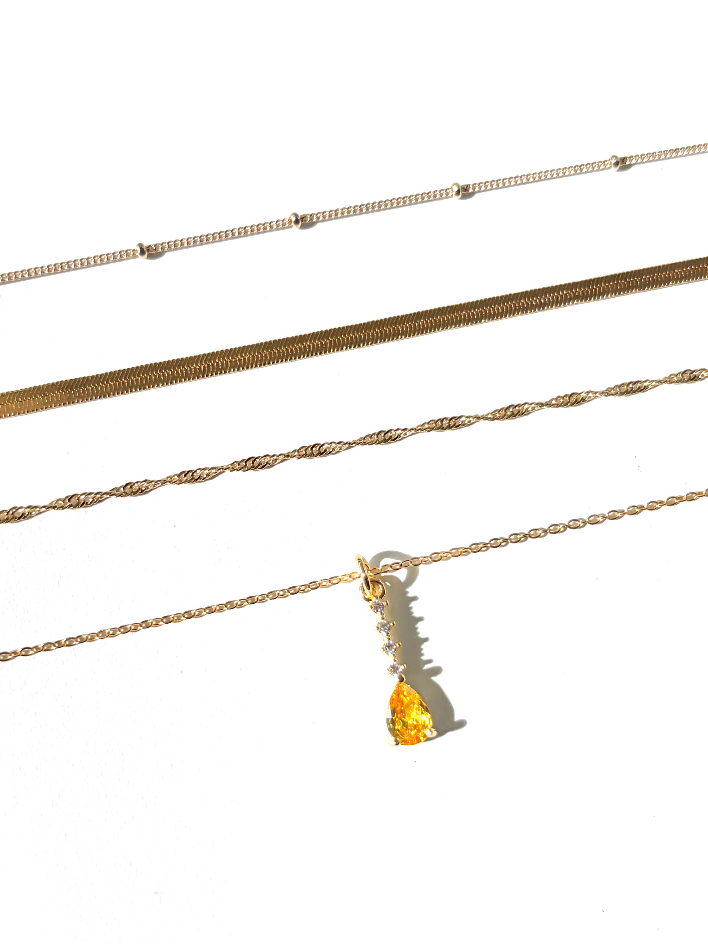 Four gold necklaces from the LLUME Jewelry Sunspill Solstice Set are displayed in parallel on a white background; one features a yellow teardrop-shaped pendant.