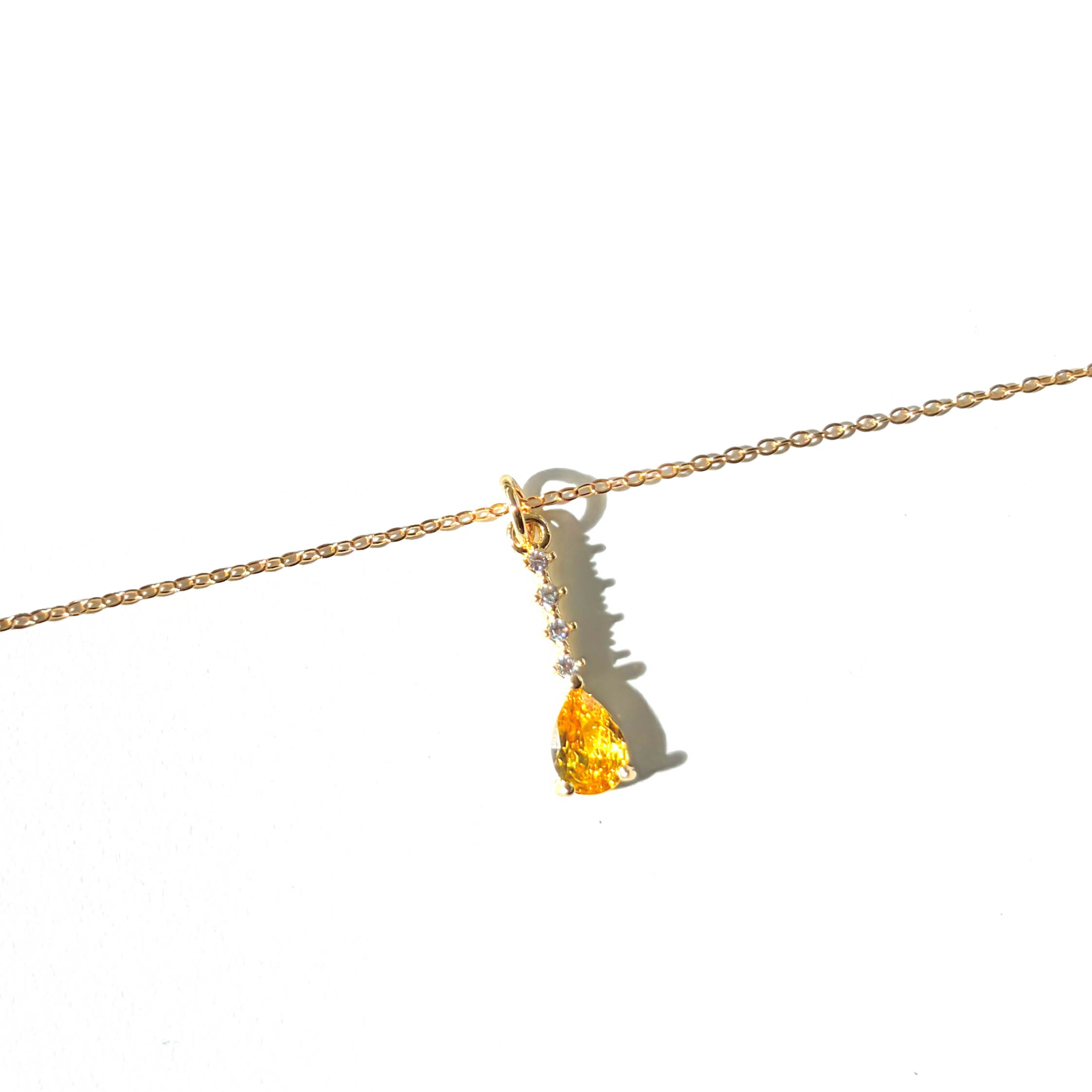 The Sunspill Necklace by LLUME Jewelry showcases a delicate gold multi-length chain with a yellow teardrop-shaped gemstone pendant, accented by small clear stones set above it, and is displayed against a white background.