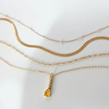 Four gold necklaces from LLUME Jewelry’s Sunspill Solstice Set are displayed on a white surface; three have simple chain designs, while one features a yellow teardrop-shaped pendant. Soft lighting casts gentle shadows.