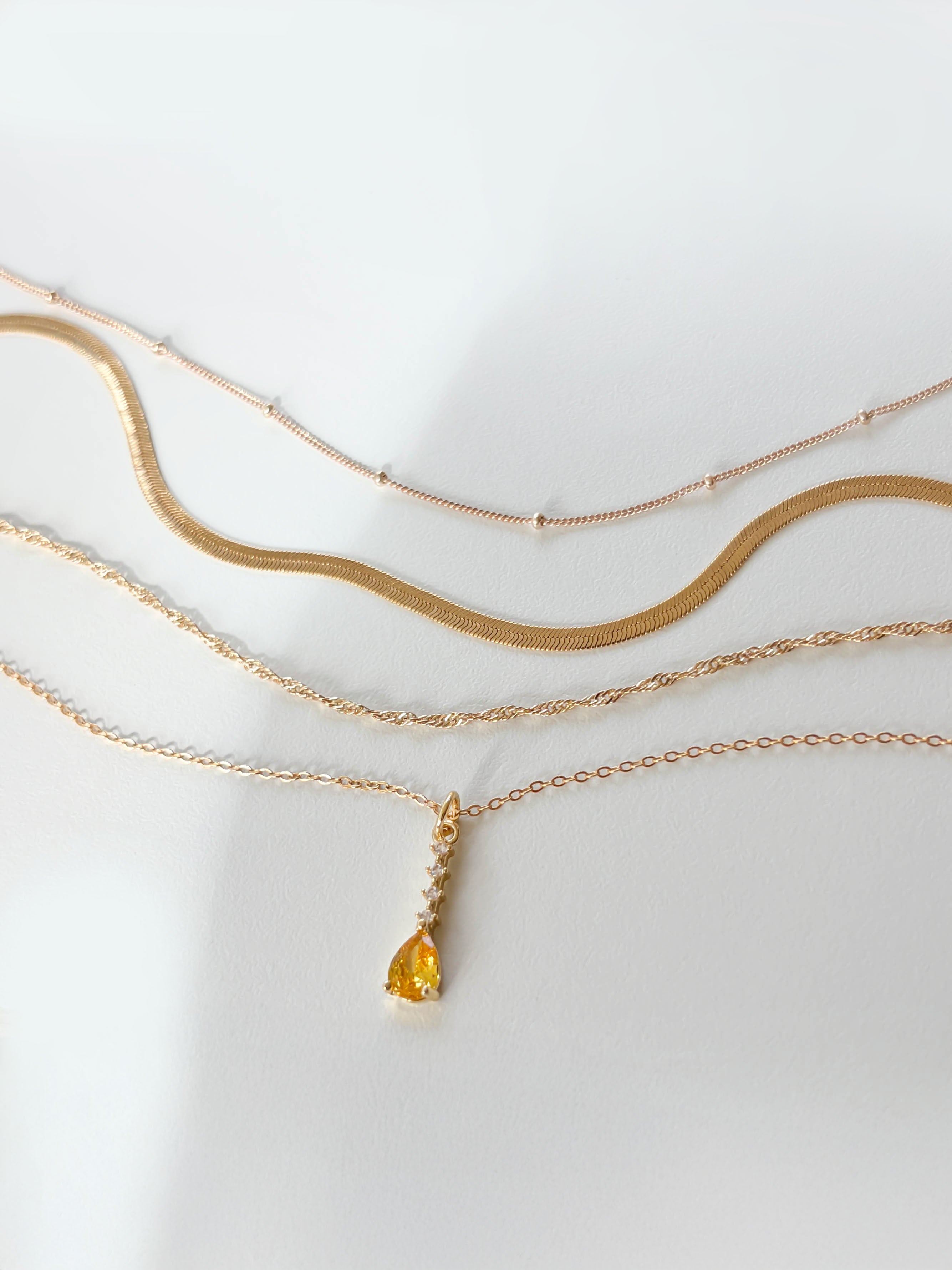 Four gold necklaces from LLUME Jewelry’s Sunspill Solstice Set are displayed on a white surface; three have simple chain designs, while one features a yellow teardrop-shaped pendant. Soft lighting casts gentle shadows.