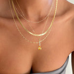 Close-up of a woman's neck and upper chest adorned with LLUME Jewelry's Sunspill Solstice Set, featuring five layered gold necklaces with a yellow gemstone pendant. She wears a grey off-shoulder top, highlighting this elegant collection.