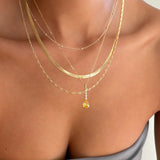 Close-up of a woman's neck and upper chest adorned with LLUME Jewelry's Sunspill Solstice Set, featuring five layered gold necklaces with a yellow gemstone pendant. She wears a grey off-shoulder top, highlighting this elegant collection.