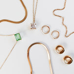 The LLUME Jewelry Vacation Capsule Set features thin gold chain necklaces—one with a green gemstone, one with a square clear stone—and pairs of gold hoop earrings, all arranged on a white surface for a sparkling display.
